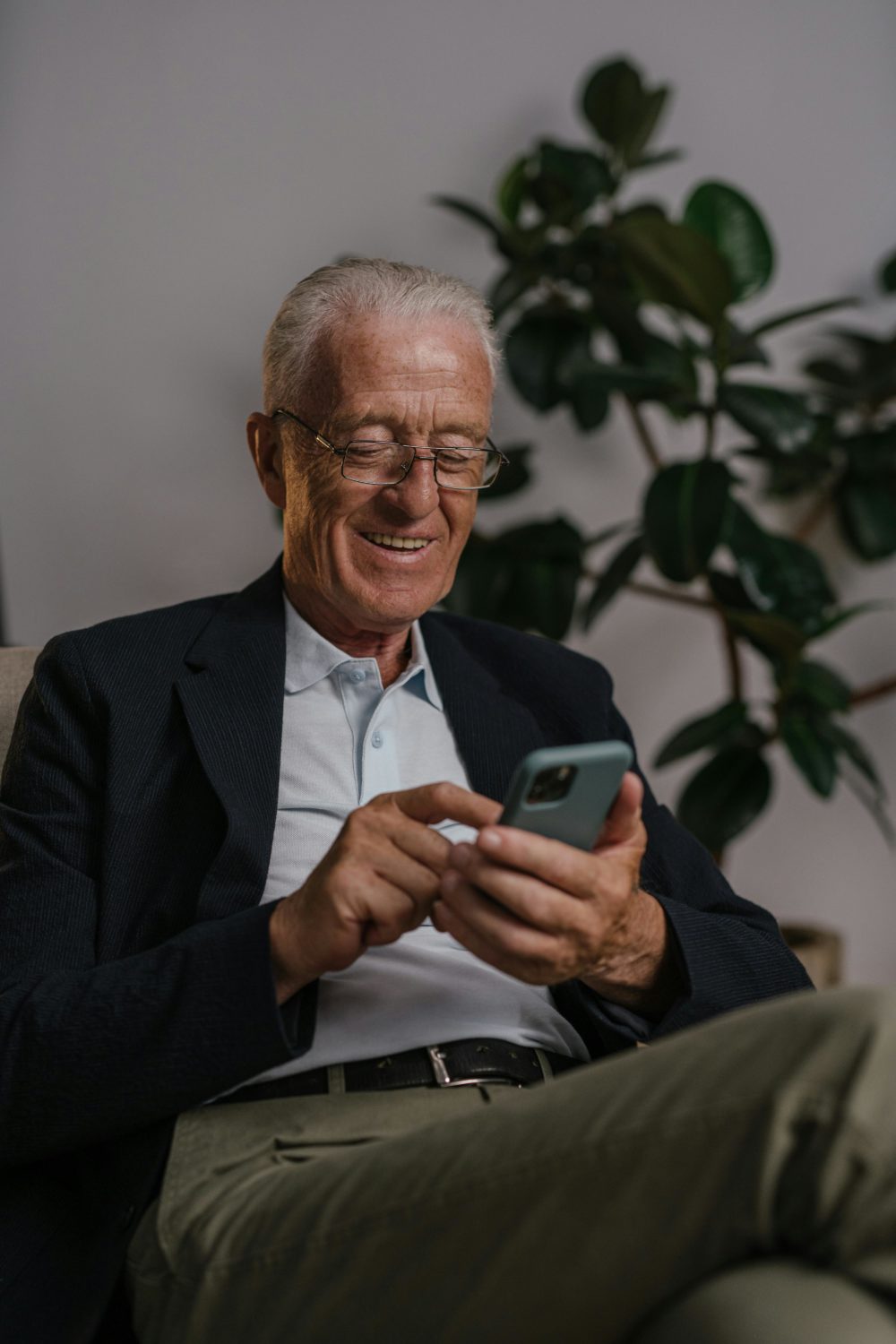 Elderly man using a smartphone, smiling, and sitting in a comfortable setting indoors.